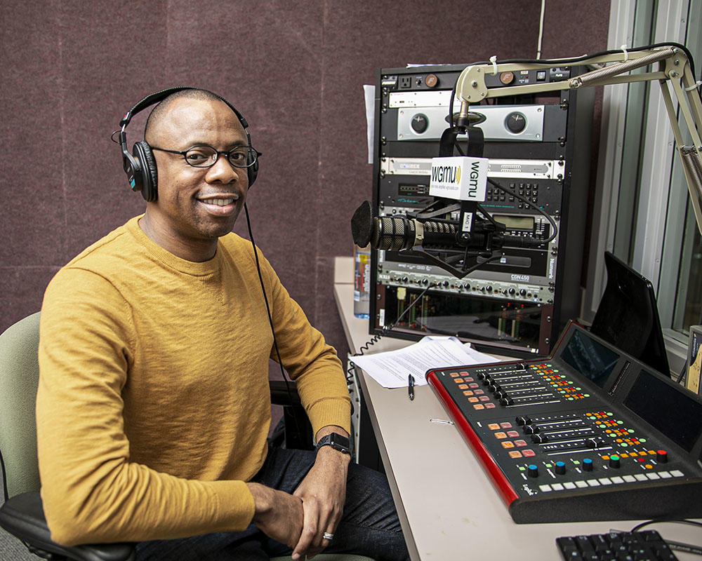 A Black man wearing an yellow long-sleeved shirt sits in a recording booth. He is wearing headphones and speaking into a mic.